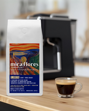 Specialty coffee bag for Max Caffeine light roast with miraflores branding and art masterpiece The Scream by Edward Munch, set on a counter with a shot of espresso and an espresso machine