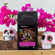 A bag of Mexican Chocolate Flavored Medium Roast coffee beans with a colorful design, accompanied by whole coffee beans and skulls on a wooden surface.