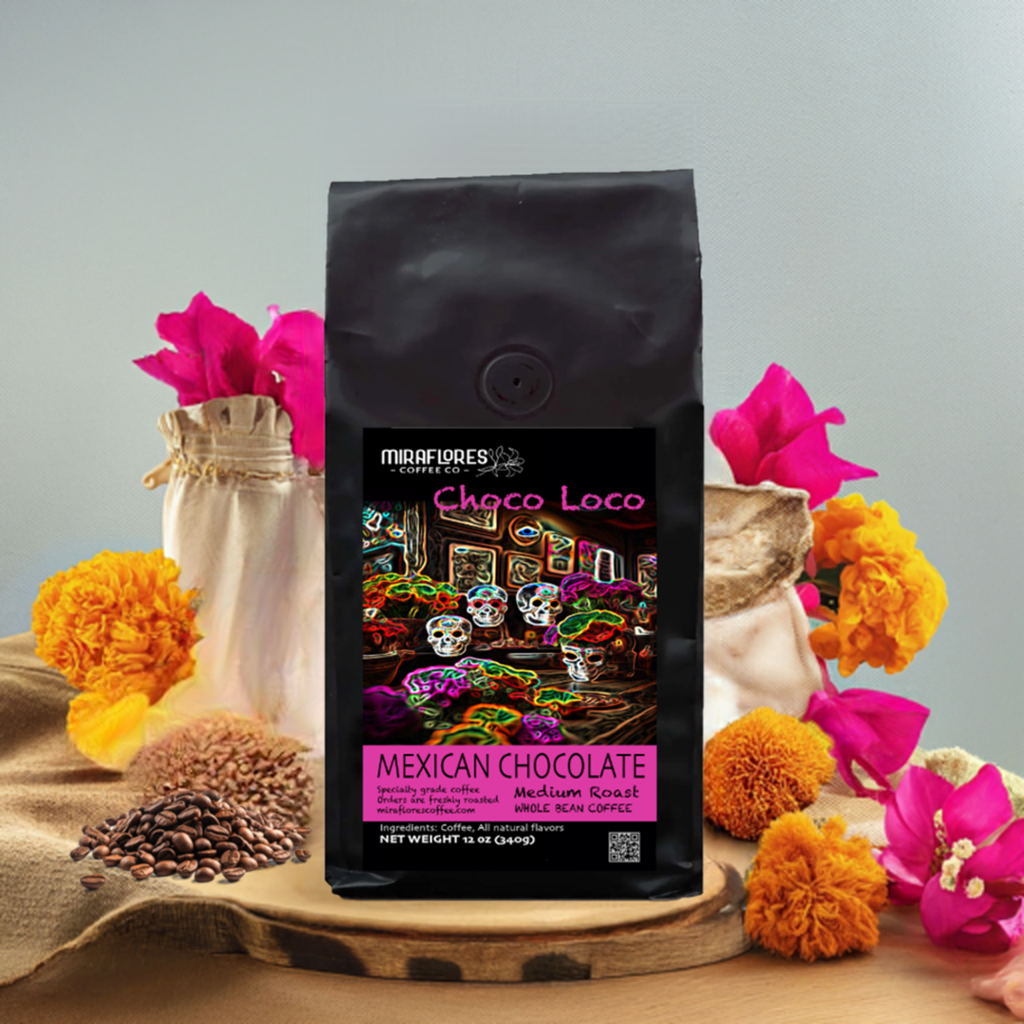 A bag of Mexican Chocolate Medium Roast coffee beans with a colorful design, accompanied by whole coffee beans and flowers on a wooden surface.