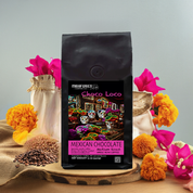 A bag of Mexican Chocolate Medium Roast coffee beans with a colorful design, accompanied by whole coffee beans and flowers on a wooden surface.