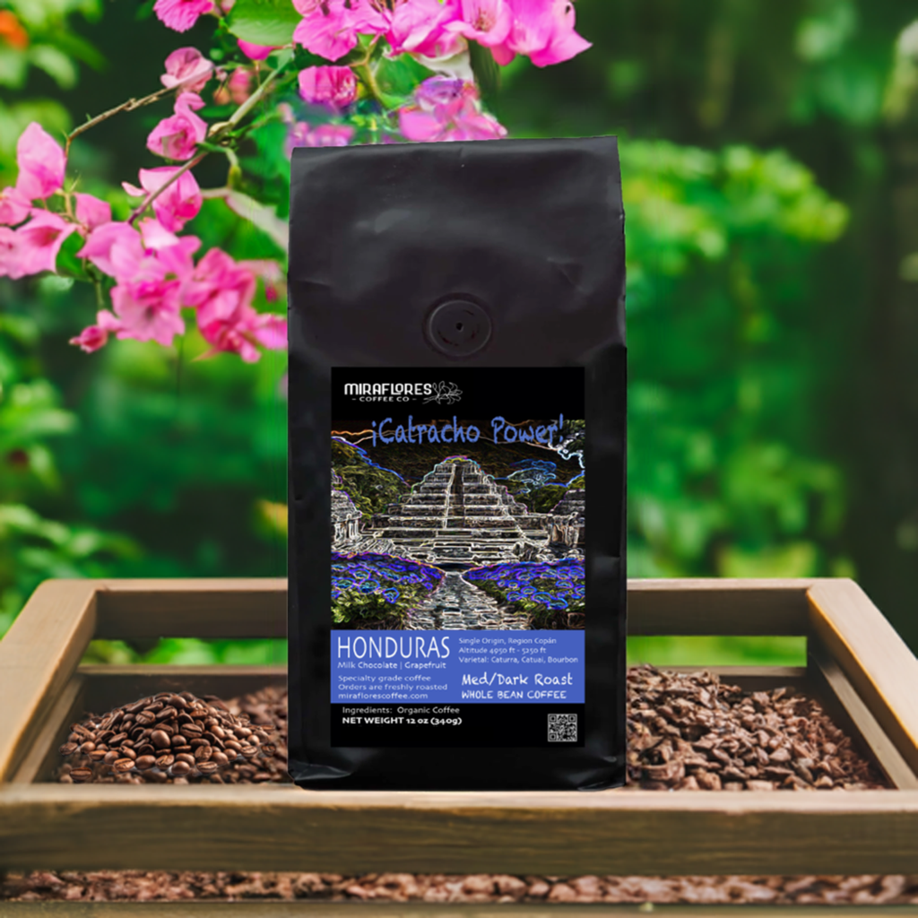 A bag of Miraflores coffee with a dark roast design, named 'Catracho Power: Honduras Medium Dark Roast', placed on a wooden tray with a background of pink flowers and green leaves.