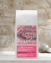 Specialty Coffee Beans bag of Mexican Chocolate flavored Coffee, Medium Roast, with pink design featuring a La Catrina by Jose Guadalupe Posada and Miraflores Coffee Co branding on a stone surface 