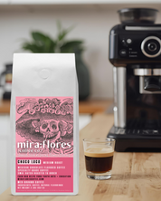 Specialty Coffee Beans bag of Mexican Chocolate flavored Coffee, Medium Roast, with pink design featuring a La Catrina by Jose Guadalupe Posada and Miraflores Coffee Co branding with a shot of espresso in a kitchen with an espresso machine