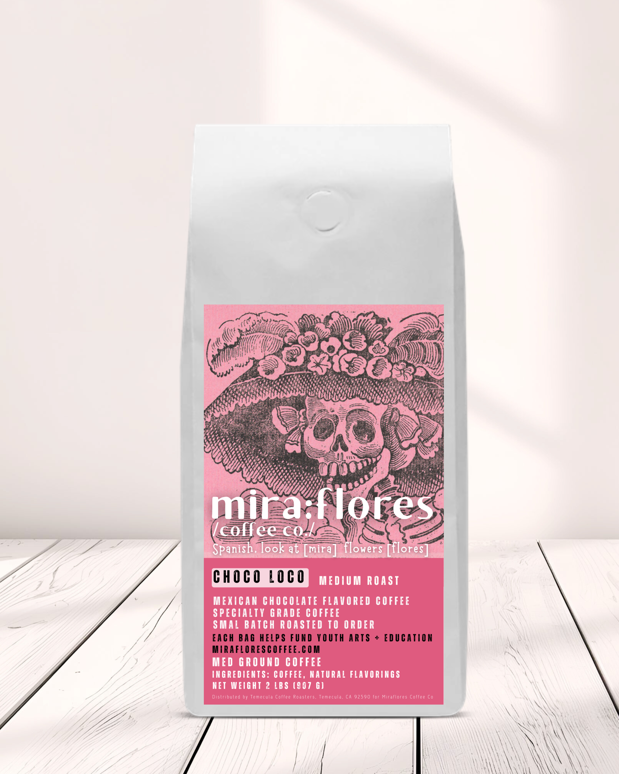 Specialty Coffee Beans bag of Mexican Chocolate flavored Coffee, Medium Roast, with pink design featuring a La Catrina by Jose Guadalupe Posada and Miraflores Coffee Co branding on a white wood counter