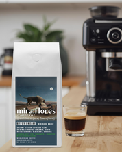 A bag of specialty coffee for a dreamy dark roast espresso made from beans from Africa sits on a counter with a shot of espresso and a coffee maker. The coffee bag has Miraflores Coffee branding and features art masterpiece The Sleeping Gypsy by Henri Rousseau.