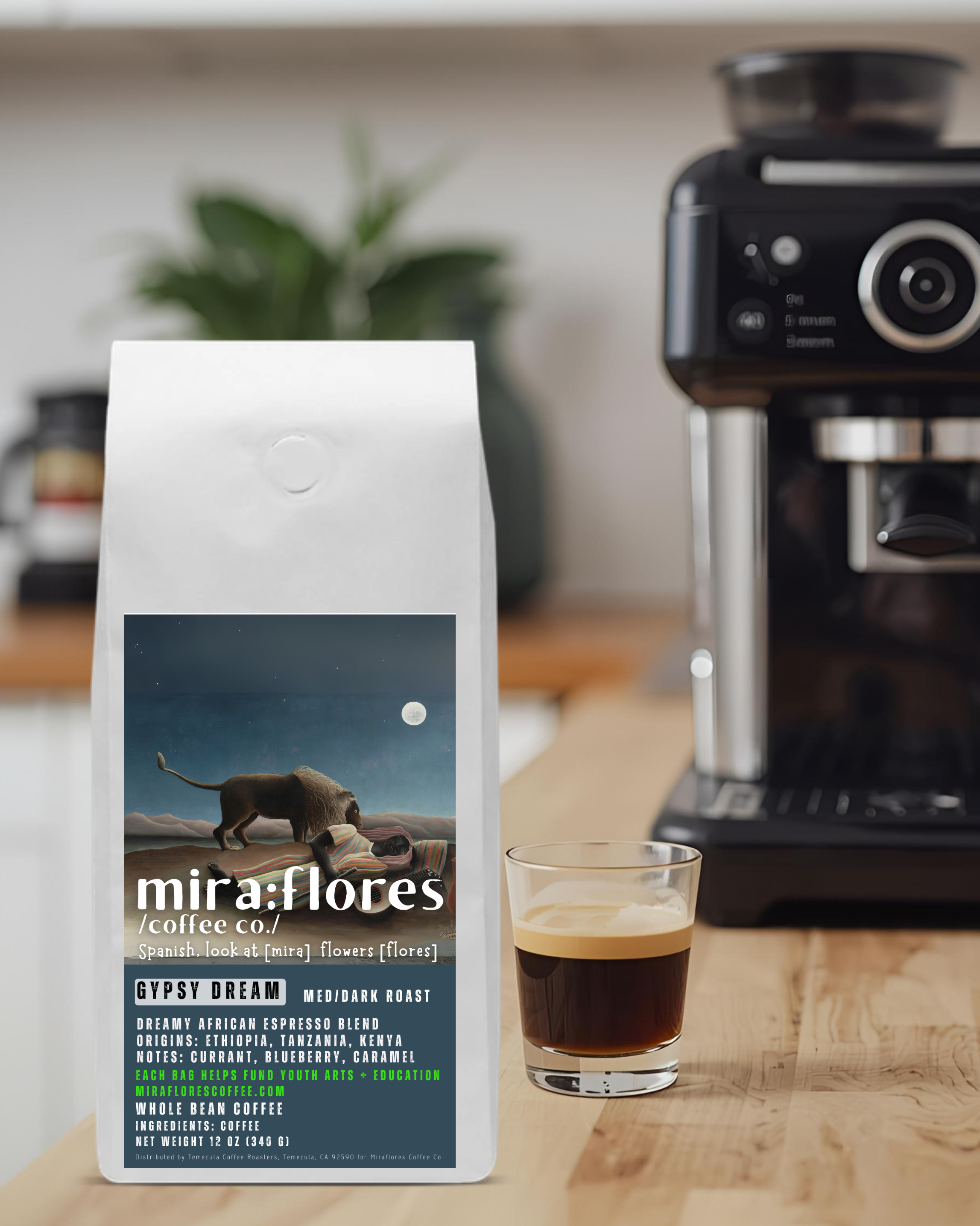 A bag of specialty coffee for a dreamy dark roast espresso made from beans from Africa sits on a counter with a shot of espresso and a coffee maker. The coffee bag has Miraflores Coffee branding and features art masterpiece The Sleeping Gypsy by Henri Rousseau.